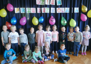 Cała grupa grupa pozuje do wspólnego zdjęcia. W tyle napis Dzień Przedszkolaka i balony ozdobione przez dzieci.